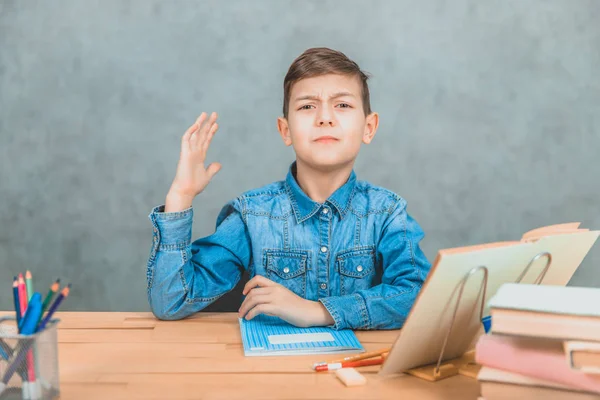 Colegial sentado en el escritorio, levantando la mano, queriendo salir. Expresión de cara divertida . — Foto de Stock