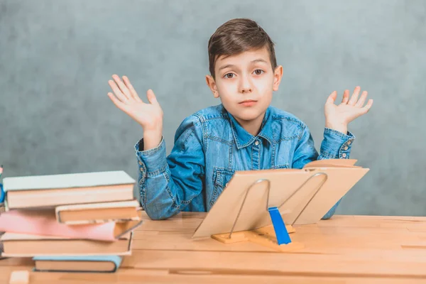 Lite trött skolpojke sitter vid skrivbordet, höja sina händer, visar att han inte vet vad de ska besvara. — Stockfoto