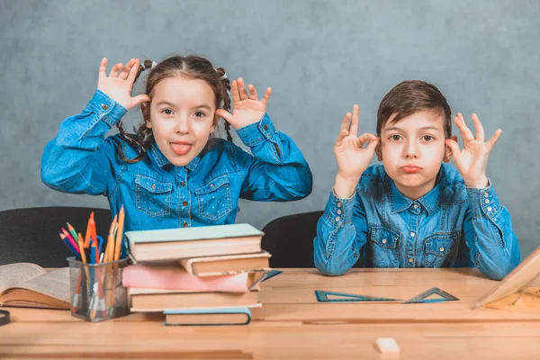 Completamente pupilos en camisetas vaqueros azules sentados en el escritorio, haciendo muecas, mostrando la lengua, levantando las manos . — Foto de Stock