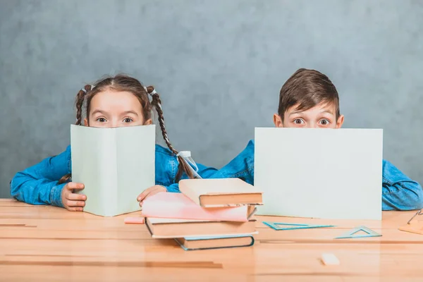 Curioso niño y niña escondidos detrás de las hojas de papel, mirando hacia arriba, a la cámara, conspiciuosly . — Foto de Stock