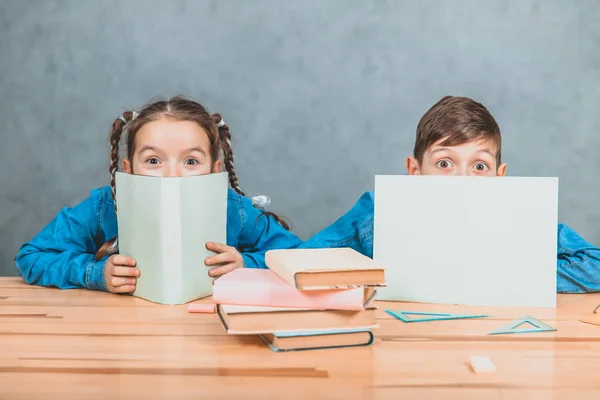 Curioso niño y niña escondidos detrás de las hojas de papel, mirando hacia arriba, a la cámara, conspiciuosly . — Foto de Stock