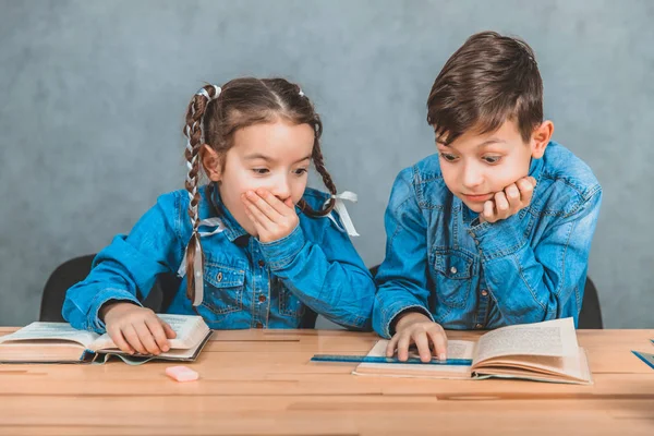 Garçon et fille intelligents totalement impliqués dans le processus de lecture. Boy pointe quelque chose d'intéressant dans son livre. Fille est surprise, couvrant sa bouche avec une main . — Photo