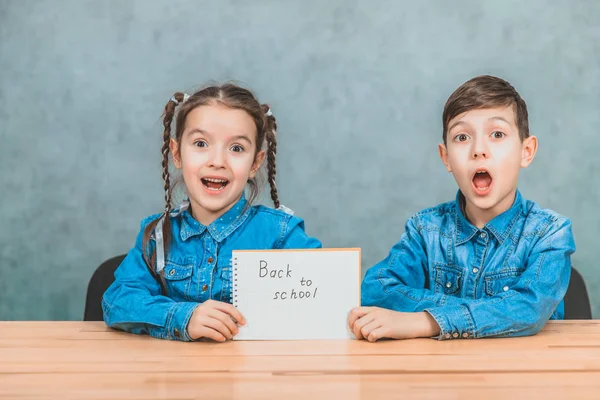 Chico y chica guapos sentados en el escritorio sosteniendo una hoja de papel con la frase de vuelta a la escuela escrita en ella. No les gusta la idea de volver a la escuela. . — Foto de Stock