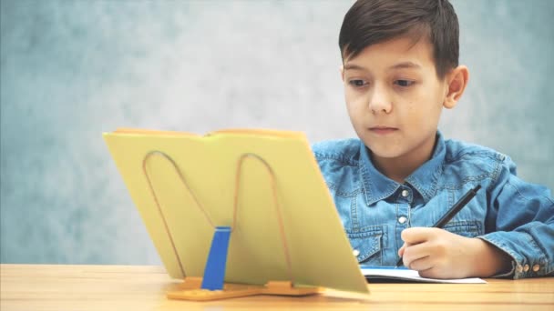 Молодой концентрированный школьник в синих джинсах пишет футболки, копируя текст из книги на книгодержателе . — стоковое видео
