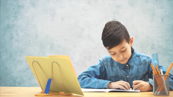 Молодой концентрированный школьник в синих джинсах пишет футболки, копируя текст из книги на книгодержателе. К нему присоединяется его сестра, которая смотрит, как он пишет. Он отгоняет ее. . — стоковое видео