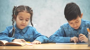 Okul çocukları, erkek ve kız kardeş, masada oturup yazıyorlar. Güzel kız ödevini yapamıyor ve yardım için kardeşi yalvarıyor.