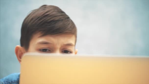 Bonito menino de escola sentado na mesa com o laptop, jogando o jogo. Expressão facial confusa . — Vídeo de Stock