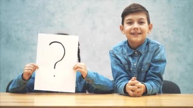Masabaşında oturan öğrenciler. Kız ünlem işareti olan kağıdın arkasına saklanıyor. Oğlan ellerini fırlatıyor, parmağını kıza fırlatıyor. Şaşkın yüz ifadesi.