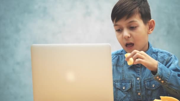 Menino bonito sentado na mesa com o laptop, pesquisando na internet e comendo uma maçã . — Vídeo de Stock