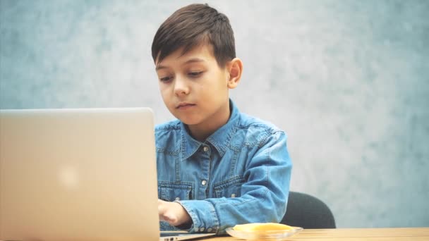 Schattige kleine school jongen zittend aan het Bureau met de laptop, zoeken in het internet en het eten van een appel. — Stockvideo