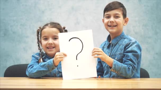 Cute schoolkids sitting at th desk, holding a sheet of paper with the exclamation mark written on it, having no idea what it means. Asking face expressions. — Stock Video