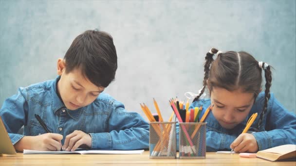 Crianças em idade escolar, irmão e irmã, sentados à mesa, escrevendo, de repente olhando um para o outro de forma conspiciosa e continuar escrevendo . — Vídeo de Stock