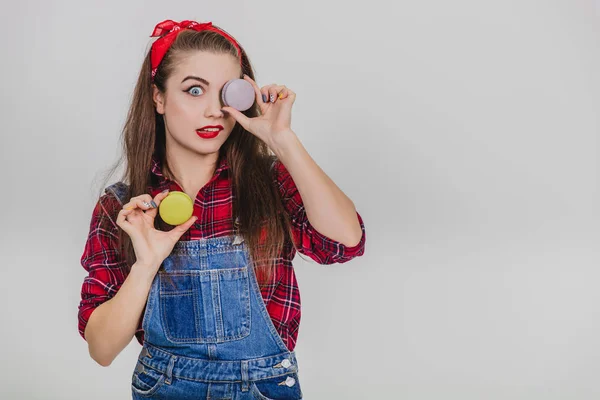 Linda feliz bonito jovem bonita mulher segurando dois macaroons, escondendo seu olho atrás de um azul, torcendo o lábio . — Fotografia de Stock