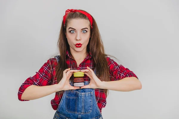 Menina engraçada agradável em pé vermelho, segurando uma pilha de saborosos macaroons coloridos em suas mãos na frente do peito, fazendo cara de pato . — Fotografia de Stock