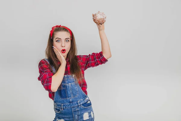 Linda jovem fêmea levantando grande donut de chocolate, mão na bochecha, satisfeito e surpreso expressão facial . — Fotografia de Stock