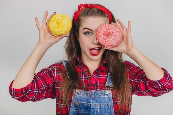 Jeune fille enfantine cache son oeil derrière un gros beignet avec du glaçage rose, tenant un beignet jaune près de la tête, la bouche largement ouverte . — Photo