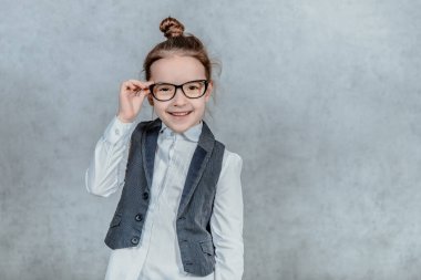 Gri arka planda gözlüklü küçük sevimli kız. Bu sırada, onun gözlüğünü tutuyor ve kameraya bakıyor..