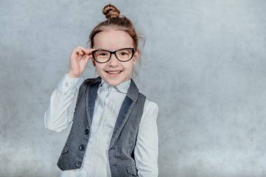 Gri arka planda gözlüklü küçük sevimli kız. Bu sırada, onun gözlüğünü tutuyor ve kameraya bakıyor..