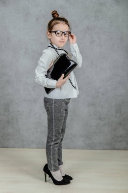 Küçük işletmeci kadının çocuğu gri bir arka planda. Elinde bir torba tutuyor. Boynunda siyah boyunları var. Gözlük takmış.