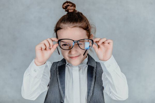 Business little cute baby girl in glasses on gray background. During this, she holds his glasses and looks at the camera. Close up.