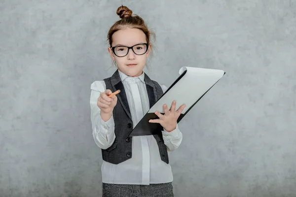 Little girl with a folder in her hands on a gray background. Dressed up as a business lady and glasses. Looking at the camera, bewildered, showing a pencil to the camera. — Stock Photo, Image
