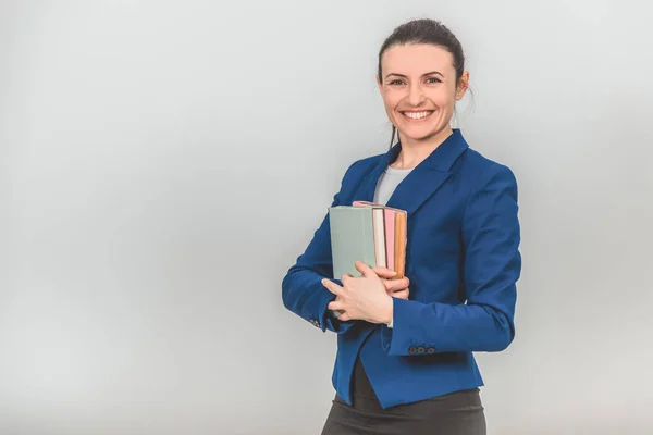 Preciosa maestra sosteniendo una pila de libros coloridos cerca de su pecho, mirando a la cámara, sonriendo . — Foto de Stock