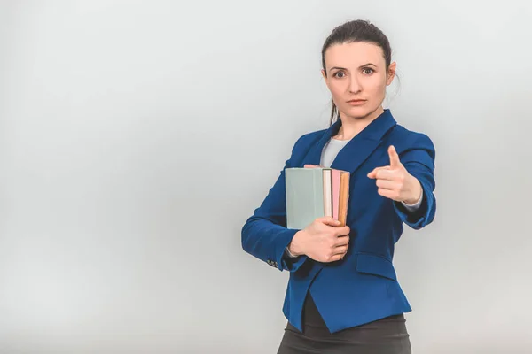 Lovely female teacher holding a stack of colorful books close to her chest, pointing her finger at the camera. — Stock Photo, Image