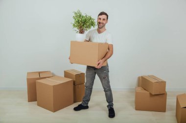 Yakışıklı genç adam yeni eve taşınıyor, ayakta, elinde karton kutu ve saksı, gülümsüyor, kutularla çevrili..