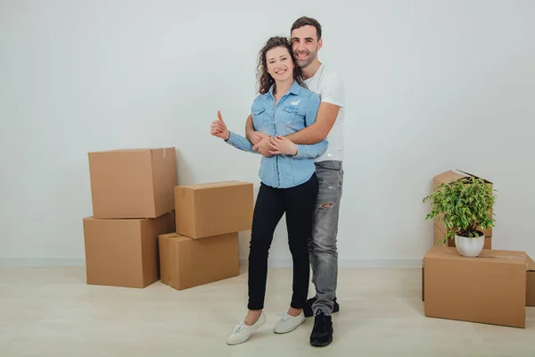 Couple moving into their new house, husband is hugging his wife, looking at the camera, smiling. Wife is giving thumb up. Room full of carton boxes. — Stock Photo, Image