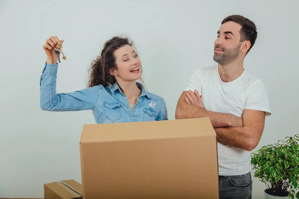 Young wife and husband standing behind the big carton box, together. Woman is holding keys to their new apartment. Man is looking at her folding his hands. — Stock Photo, Image