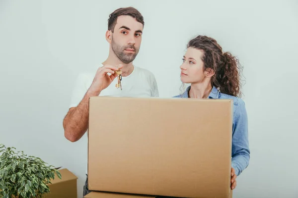 Jovem esposa e marido de pé atrás da pilha de caixas de papelão grandes, juntos. Homem está segurando as chaves de seu novo apartamento . — Fotografia de Stock