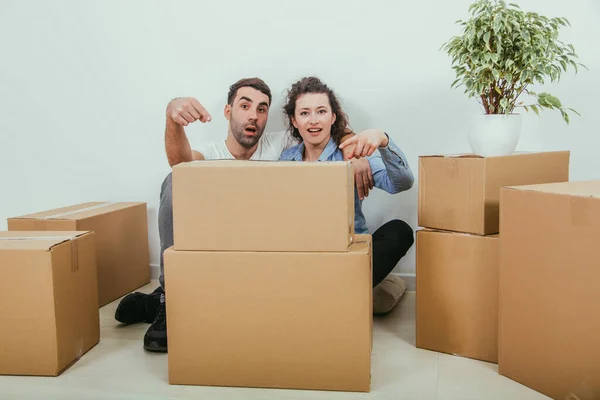 Esposa engraçada e marido brincando, sentado atrás das caixas de papelão grandes, apontando os dedos para eles, olhando tolamente . — Fotografia de Stock