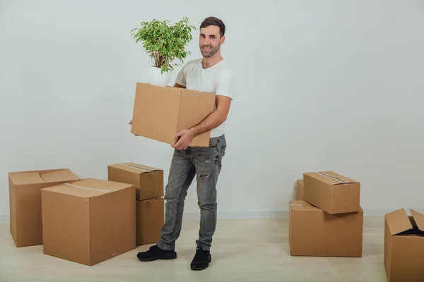 Bonito jovem movendo-se para casa nova, de pé, segurando caixa de papelão e vaso de flores, sorrindo, cercado por caixas . — Fotografia de Stock