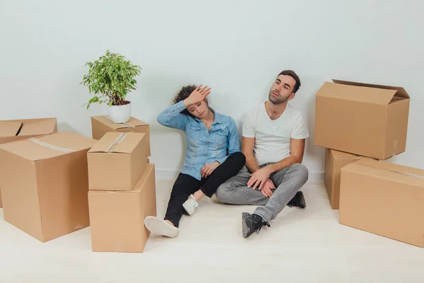 O processo de mudança para uma nova casa é totalmente cansativo. Lovely jovem casal esposa e marido estão sentados no chão em novo apartamento entre caixas de papelão . — Fotografia de Stock