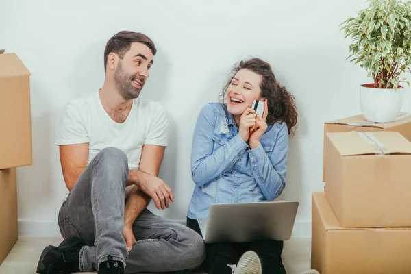 Casal jovem com laptop no apartamento, sentado no chão, surfando na internet, à procura de novos móveis. Mulher é extremamente feliz, segurando um cartão de crédito . — Fotografia de Stock