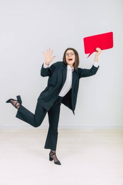 Uzun boylu, heyecanlı genç bir iş kadını tek ayak üzerinde duruyor, boş bir kare kırmızı konuşma balonunu kaldırıyor, kameraya bakıyor, gülümsüyor..