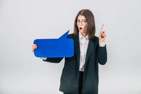 Portrait of an excited young business woman holding empty square blue speech bubble, pointing her finger up, asking to pay attention. — Stock Photo, Image