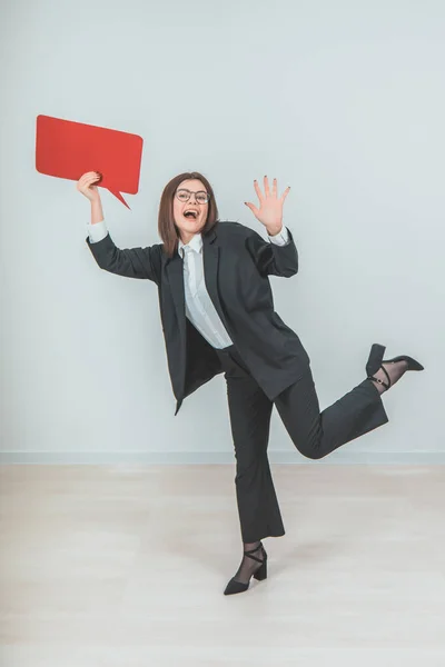 Full-length excited young business woman standing on one leg, raising up empty square red speech bubble, looking at the camera, smiling. — Stock Photo, Image