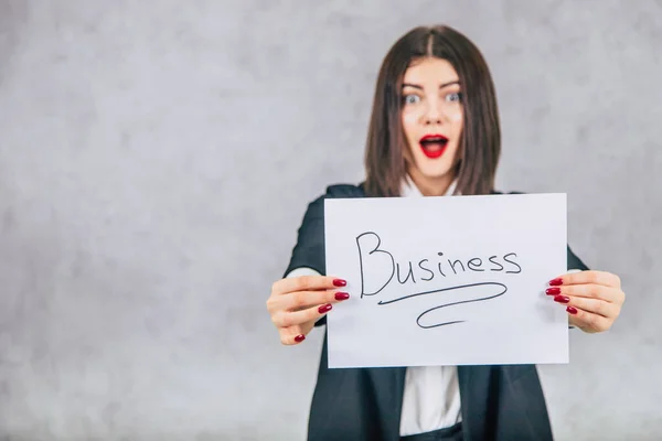 Blurred lovely businesswoman in black suit standing, extending to the camera a sheet of paper with word business written on it. Focus on the paper. — Stock Photo, Image