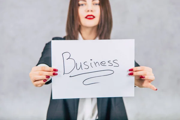 Blurred lovely businesswoman in black suit standing, extending to the camera a sheet of paper with word business written on it. Focus on the paper. — Stock Photo, Image