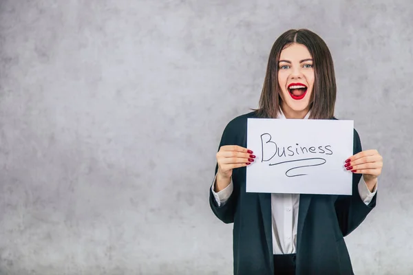 Femme d'affaires inspirée en costume noir debout avec presse-papiers dans les mains, souriante, la bouche largement ouverte. Le mot business est écrit sur la feuille de papier . — Photo