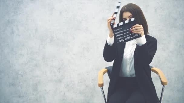 Cute young girl sitting on the chair, clapping a clapperboard in front of her face, posing, smiling. — Stock Video