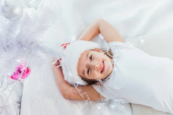 Foto von dem süßen kleinen Baby in der Weihnachtsmütze, das auf dem Bett liegt, mit Girlanden spielt, die am Tannenzweig hängen, lieblich lächelt. — Stockfoto