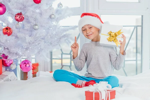 Смішний хлопчик в капелюсі Санта сидить на ліжку в положенні лотоса, тримаючи подарункову коробку і кидаючи палець вгору, звертаючи увагу на те, про що він говорить . — стокове фото