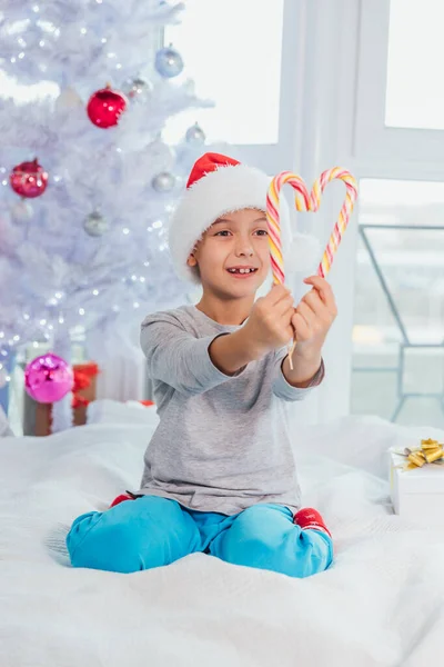 Drôle garçon dans santa chapeau est assis sur le lit en pyjama, démontrant cannes de bonbons rayés . — Photo