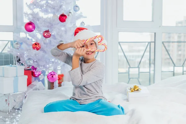 Portrait de heureux garçon préadolescent en chapeau rouge Père Noël regardant à travers le cœur de cannes de bonbons de Noël, tenant des bonbons dans ses mains devant le visage . — Photo