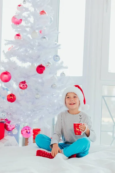 Somnolent garçon boit du cacao, assis sur le lit près de l'arbre de Noël le matin après le Nouvel An . — Photo