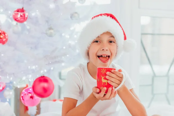 Niño guapo vestido como mini Santa o elfo está sentado cerca del árbol de Navidad con una taza de chocolate caliente . — Foto de Stock