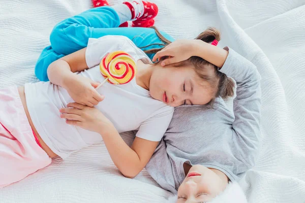 Encantadora hermana está durmiendo sobre el vientre de su hermano, sosteniendo una gran piruleta en un palo, ambos se ven dulces . —  Fotos de Stock