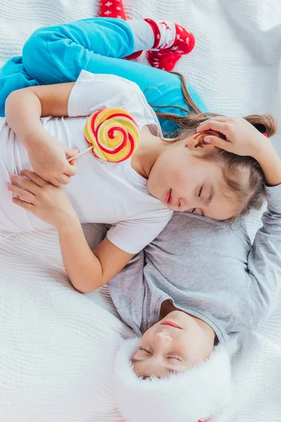 Encantadora hermana está durmiendo sobre el vientre de su hermano, sosteniendo una gran piruleta en un palo, ambos se ven dulces . — Foto de Stock